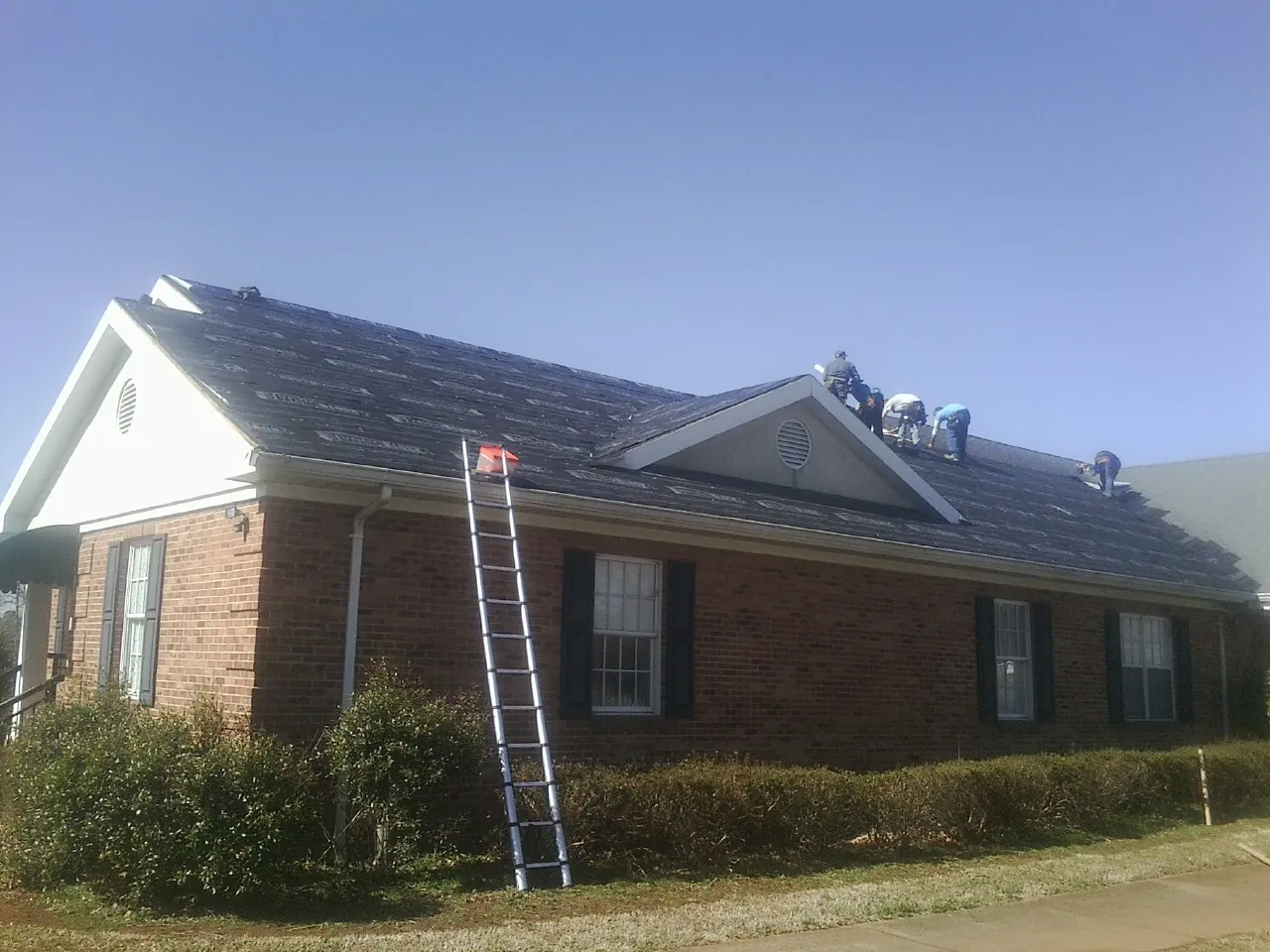 Crew tearing shingles off a brick ranch roof with a ladder set — Metal Roof Repair work by our crew in Godwin